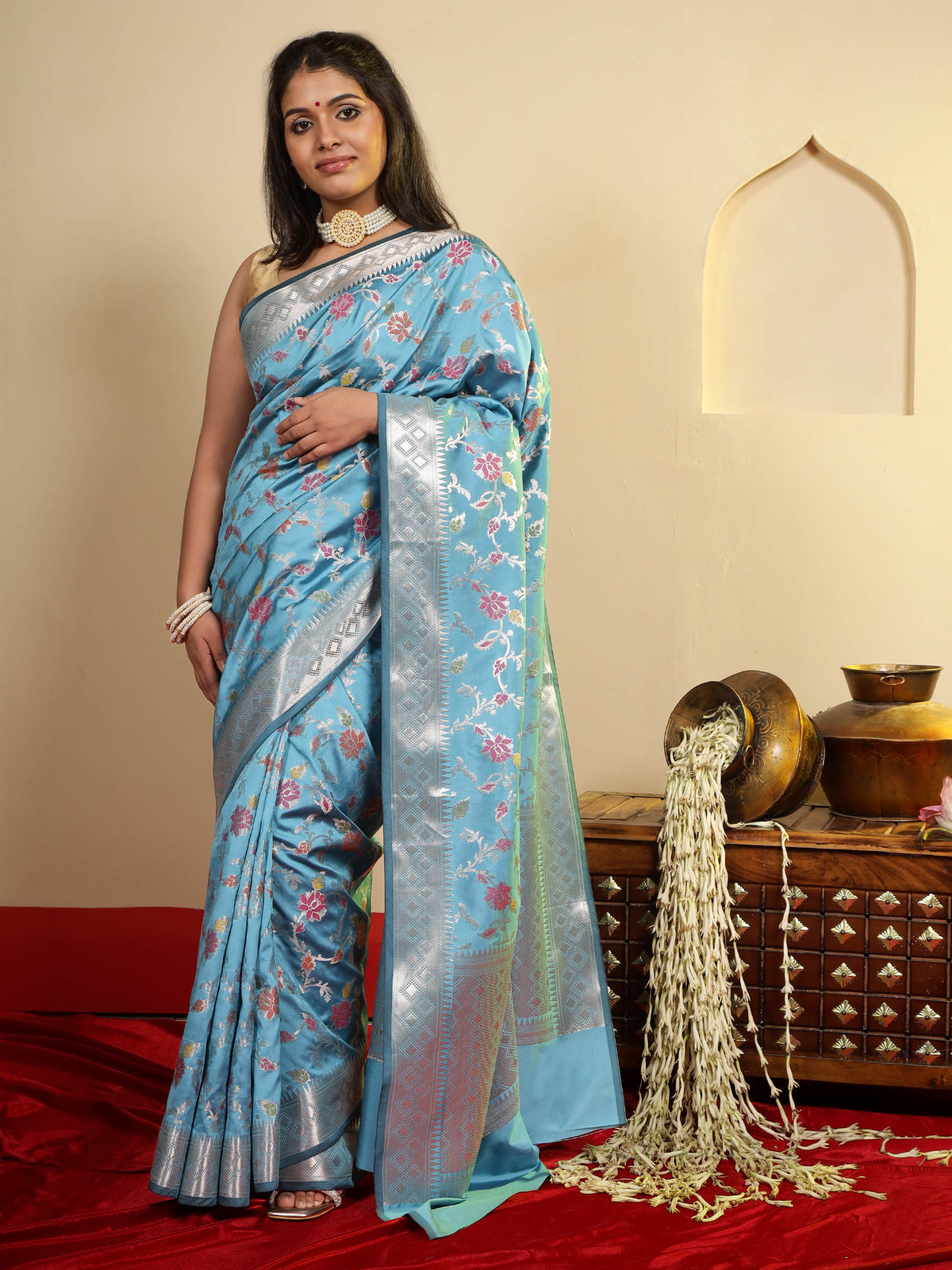 A woman modeling a light blue silk saree with silver zari embroidery and pink floral motifs, standing in a decorated room with a red carpet and traditional decor.